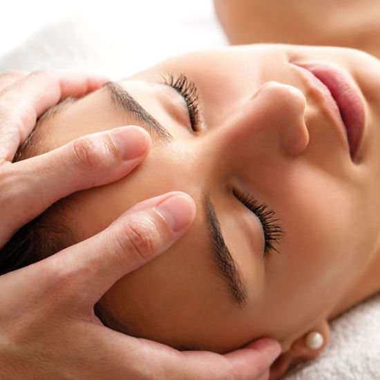 Das Bild zeigt eine Nahaufnahme einer Person, die eine Gesichtsmassage erhält. Die Augen sind geschlossen, während eine andere Person mit den Händen sanft das Gesicht massiert. Die Szene wirkt entspannend und erinnert an eine Spa- oder Kosmetikbehandlung mit ruhiger, beruhigender Atmosphäre.