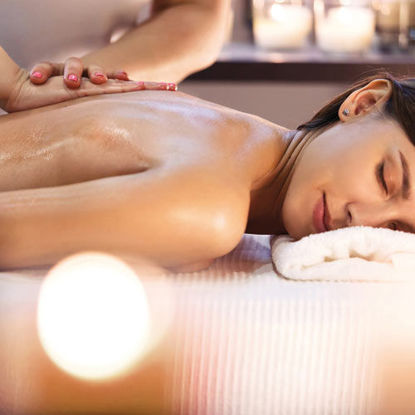 Eine Frau liegt entspannt auf einer Massageliege, während ihr Rücken von einer Masseurin sanft eingeölt und massiert wird. Ihr Kopf ruht auf einem weißen Handtuch, die Augen sind geschlossen. Im Hintergrund sorgen Kerzen für eine stimmungsvolle Wellness-Atmosphäre.