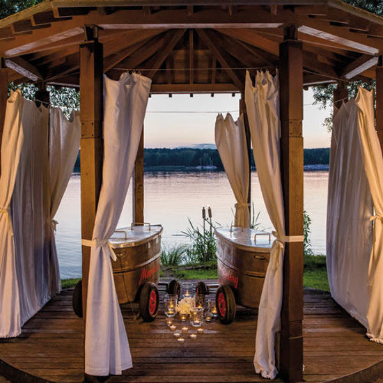 Romantischer Pavillon mit weißen Vorhängen und zwei nostalgischen Badewannen auf Holzplattform, beleuchtet von Kerzenlicht mit Blick auf einen ruhigen See bei Sonnenuntergang.